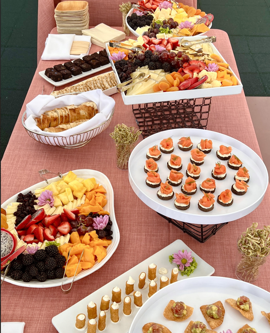 A beautifully arranged catering spread by Sweet Basil Catering, featuring fresh fruit platters, smoked salmon canapés, mini desserts, and bite-sized hors d'oeuvres at a social event.